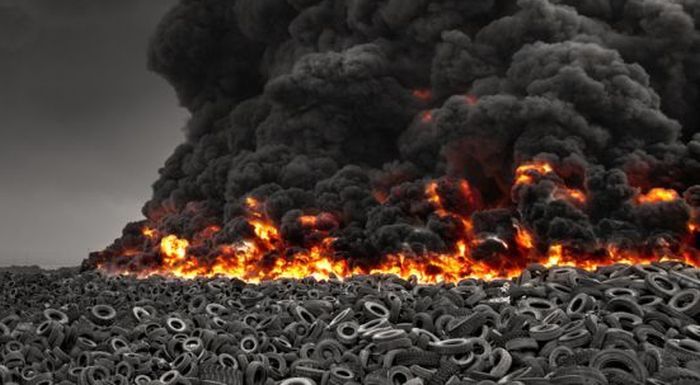 Large fire burning across a pile of discarded tires, producing thick black smoke and orange flames against a gray sky.