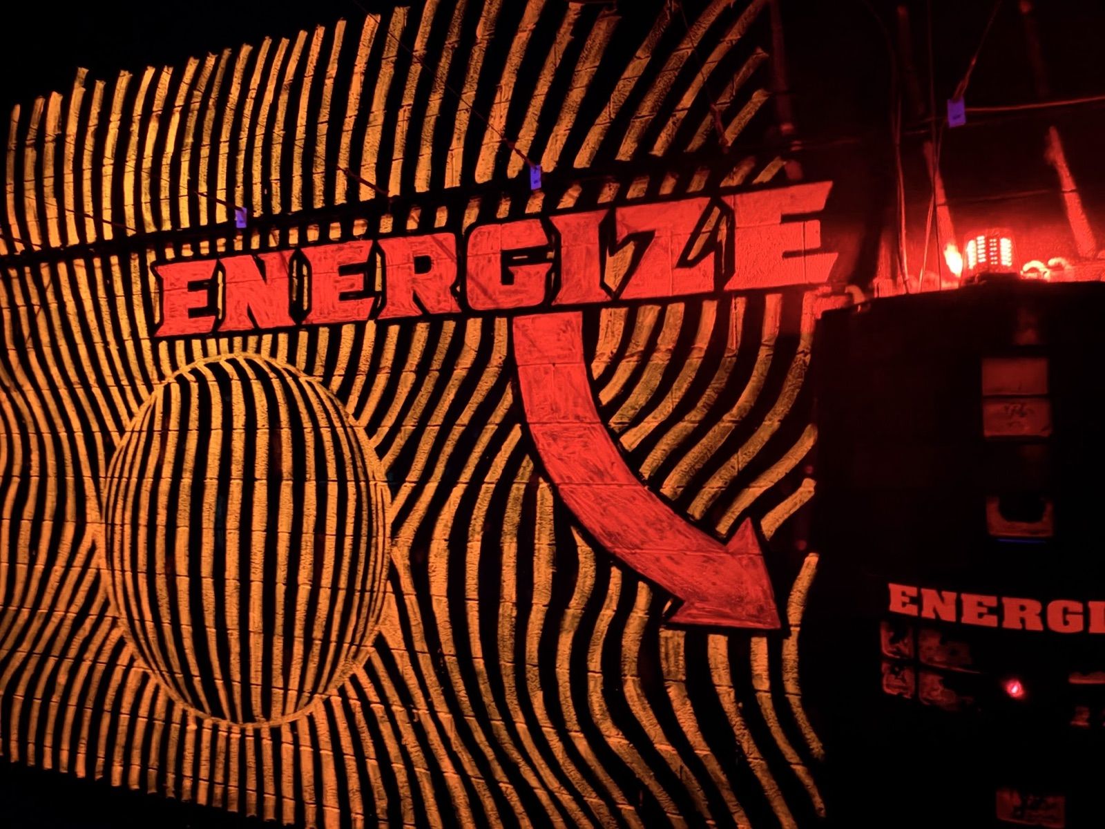 A wall of warped orange and black stripes with a big red "ENERGIZE" sign and arrow pointing to a charging station