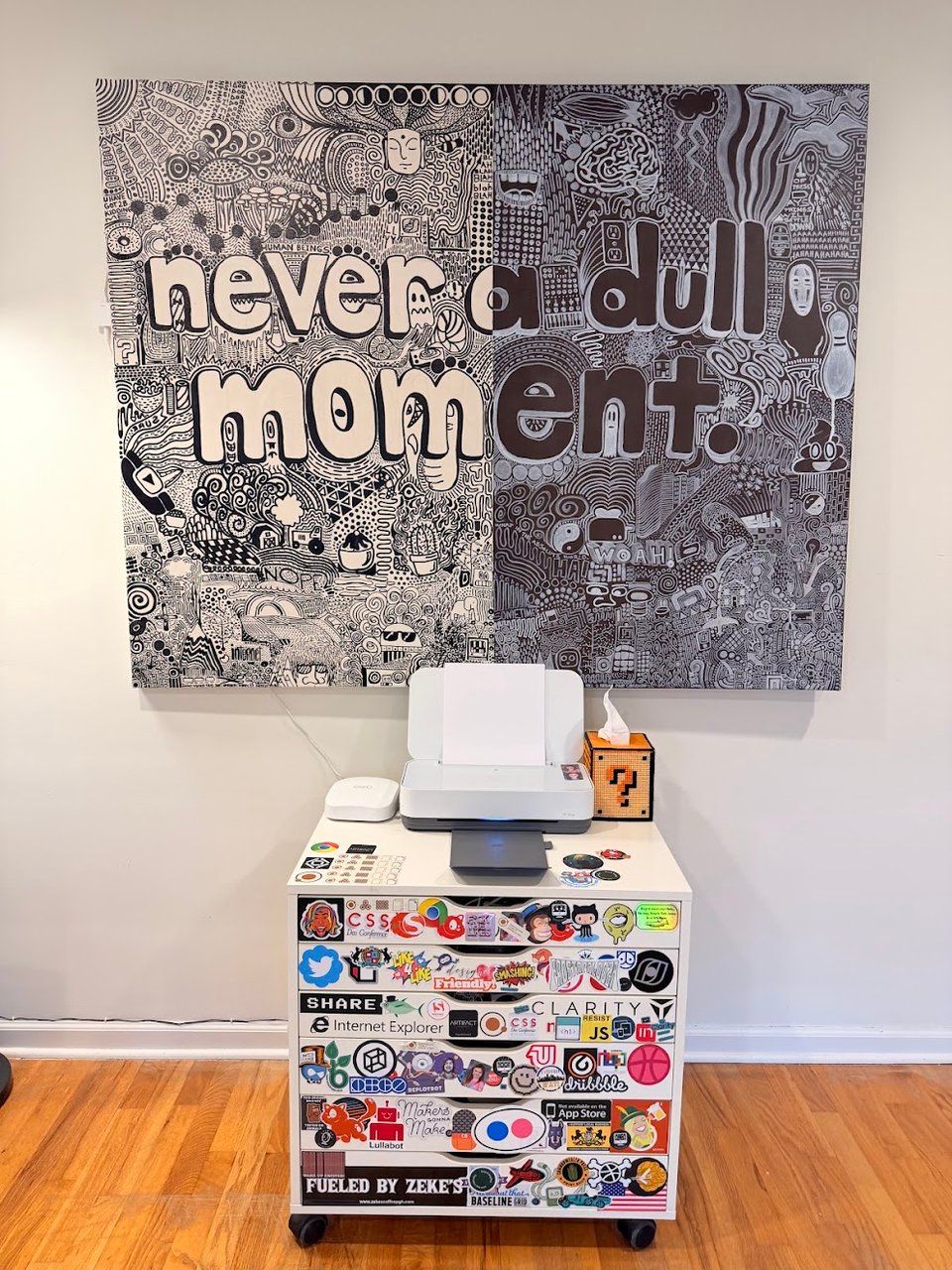 A large black and white intricate line drawing painting with text reading "never a dull moment". Beneath it is an Ikea storage cart covered with sticker labels, supporting a printer and tissue box.