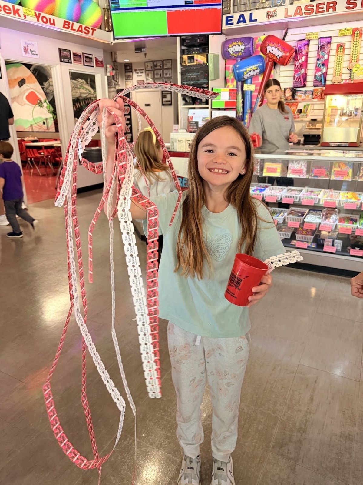 A grinning kid holding up long, tangled strands of prize tickets in one hand and a red cup of tokens in the other