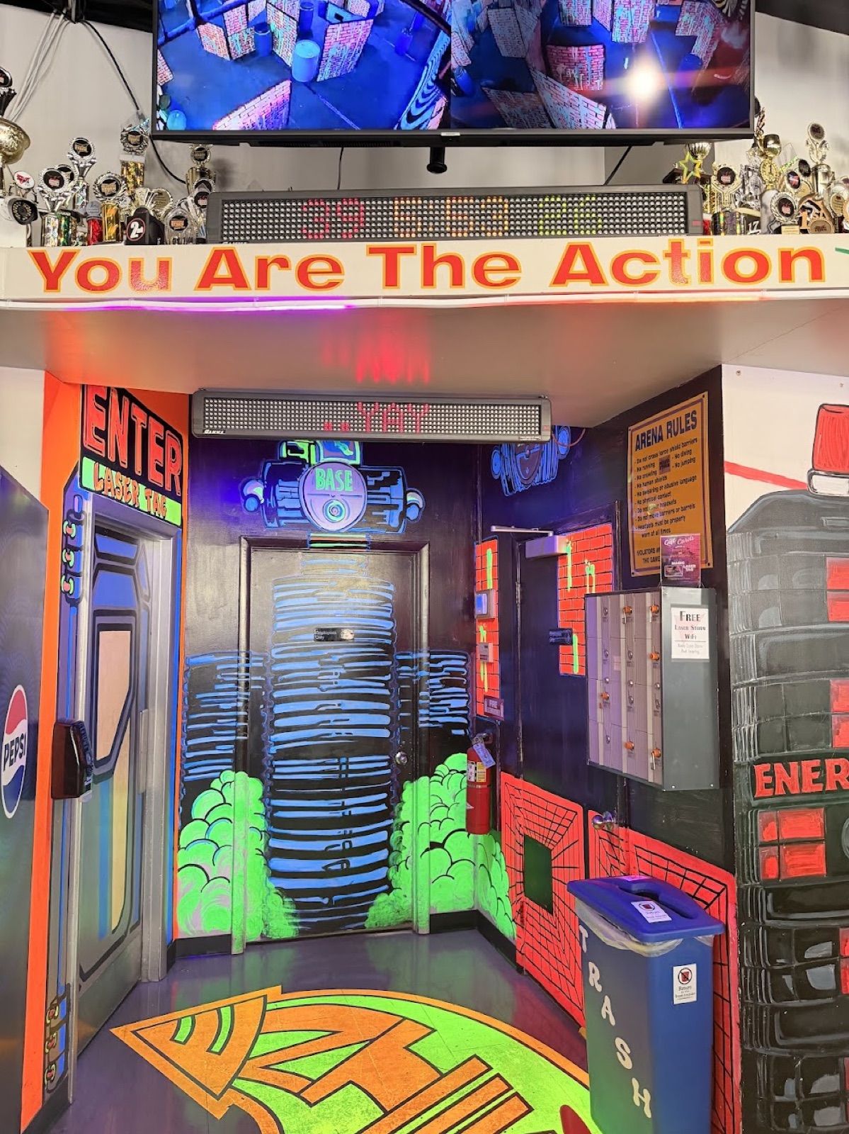 The laser tag arena entrance: a neon-painted vestibule under a "You Are The Action" banner, with trophies lining the top shelf and a monitor showing the arena