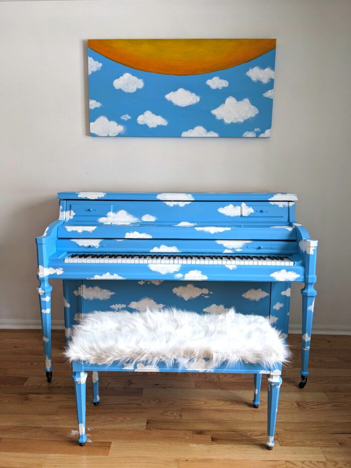 A sky blue piano with cloud pattern painted on it. A canvas with matching pattern and a semi-circle sun shape sits above the piano. A piano bench with 
