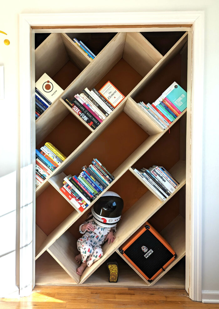 Slanted shelves in a closet with various books on them. A little girl wearing a toy space helmet sits in one of the shelves.