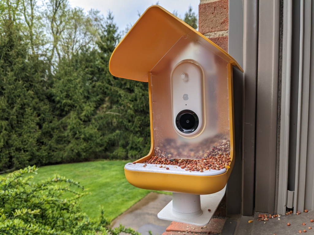 The Bird Buddy with yellow casing with white camera mounted to a window