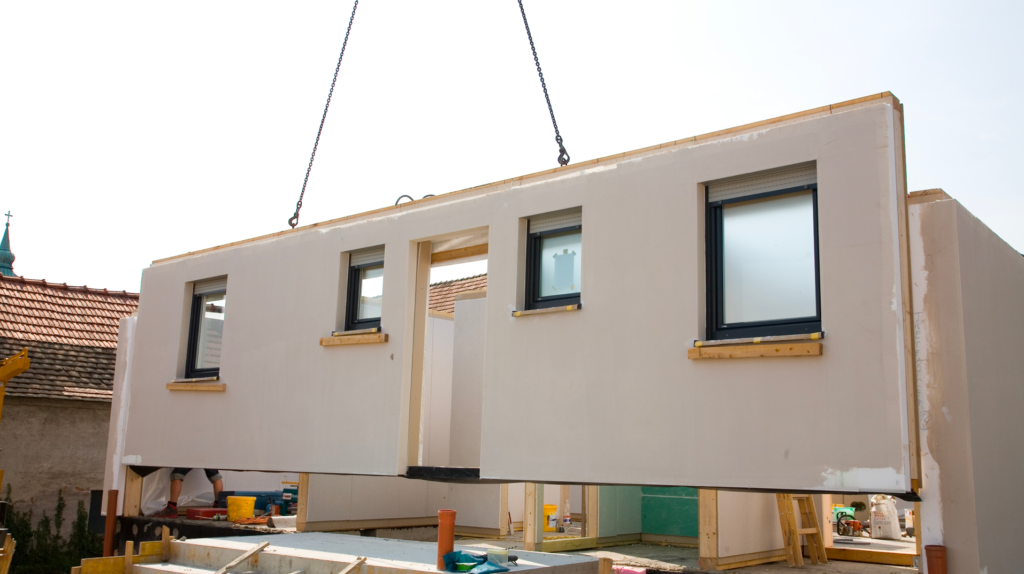 A gray prefabricated wall of a house featuring a door and 4 windows being lowered into place by a crane.